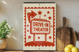 a vintage-style sign that reads "DRIVE-IN THEATER" with a red and white design, placed on a shelf next to a wooden cutting board and some potted plants.