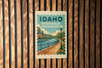A vintage-style sign for Idaho Riverwalk promenade, featuring a scenic illustration of a river, park, and street lamp.
