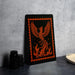 a framed artwork depicting a phoenix, a mythical bird, with flames and a fiery design on its wings. The artwork is displayed on a black surface, with a stack of black camera lenses and a white dish visible in the background.