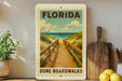 A framed poster of a wooden boardwalk leading to the beach, with the text "FLORIDA DUNE BOARDWALKS" printed on it. The poster is displayed on a wall, with a potted plant, a cutting board, and some lemons visible in the foreground.