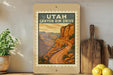 A framed poster of the scenic Utah Canyon Rim Drive, featuring a winding road and a mountainous landscape, is displayed on a shelf next to kitchen utensils and a potted plant.