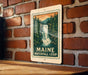 A metal sign with a waterfall and forest scene is mounted on a brick wall.