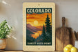 A framed vintage-style poster of the sunset over the mountains of Colorado, with the text "SUNSET VISTA POINT" printed on it. The poster is displayed on a shelf next to a wooden cutting board and a potted plant.