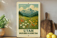 A vintage-style sign depicting a scenic mountain landscape with wildflowers, mounted on a wall next to a wooden cutting board and a potted plant.