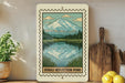 a framed illustration of a mountain lake with a wooden paddle and a wooden cutting board on a shelf.
