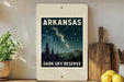 A framed poster of a night sky with stars and silhouetted trees, depicting the Arkansas Dark Sky Reserve, is displayed on a shelf next to a wooden cutting board and a potted plant.