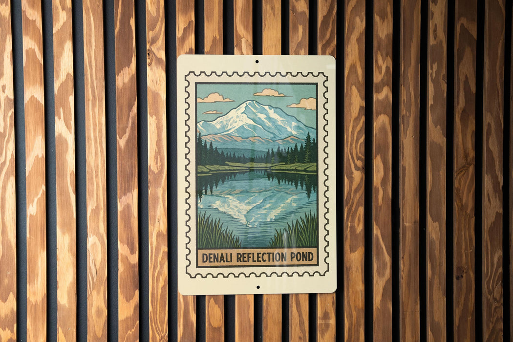 A sign for the Denali Reflection Pond, featuring a mountain landscape with a lake and trees, mounted on a wooden wall.