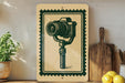 a framed illustration of a camera on a shelf, with a potted plant and a wooden cutting board visible in the background.