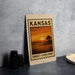 A Kansas sunset stamp or sign with a landscape image of a sunset over a field, placed on a countertop next to some black camera lenses.