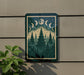 a framed artwork depicting a forest scene with pine trees and a crescent moon, mounted on a wall.