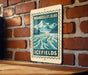 A vintage-style metal sign with a mountain landscape and a river, mounted on a brick wall.