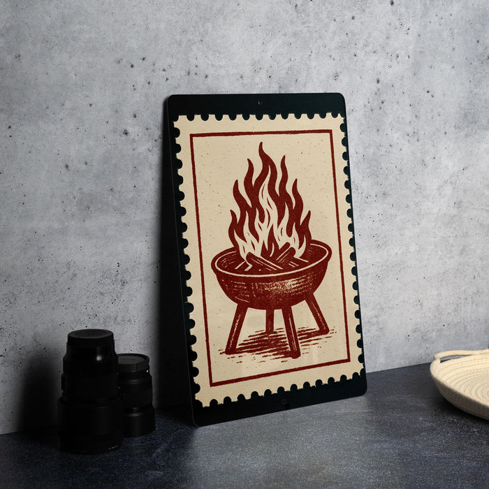 a framed illustration of a grill with flames on it, placed on a table next to some black containers.