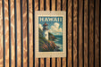 A vintage-style stamp depicting a lighthouse on a cliff overlooking the ocean, with the word "HAWAII" written on it, is displayed on a wooden wall.