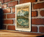 A metal sign with a mountain landscape and a lake, mounted on a brick wall.
