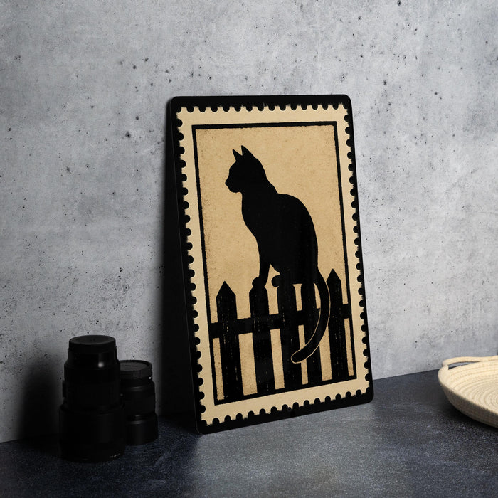 A framed black silhouette of a cat sitting on a wooden fence is displayed on a dark surface, accompanied by a stack of black camera lenses and a woven basket.