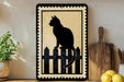 A black silhouette of a cat sitting on a wooden fence is displayed on a wall, accompanied by a potted plant, a wooden cutting board, and some lemons.