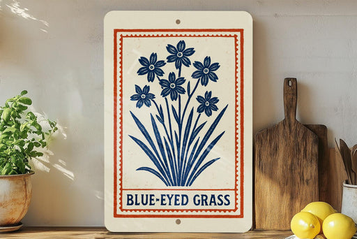 a white sign with a blue design, displaying the text "BLUE-EYED GRASS" and a decorative border. The sign is placed on a wooden shelf, with a potted plant to the left and a wooden cutting board to the right.