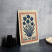 A metal sign with a blue and white design featuring a flower and the text "BLUE-EYED GRASS" is displayed on a gray countertop.