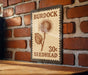 A vintage-style postage stamp featuring a thistle flower and the text "BURDOCK 30¢ SEEDHEAD" is displayed on a brick wall.