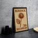 A metal sign with the text "BURDOCK 30¢ SEEDHEAD" printed on it, along with an illustration of a thistle flower. The sign is placed on a table next to some black camera lenses.