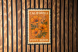 A California postage stamp featuring an illustration of orange poppies is displayed on a wooden wall.