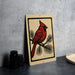 a red cardinal perched on a branch, with a beige background and a black border.