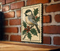 A decorative metal plaque featuring a small bird perched on a branch with berries, set against a brick wall.