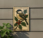 A decorative metal plaque featuring a black and white bird perched on a branch with berries, set against a gray wall.