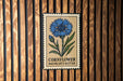 a sign with a blue flower and the text "Cornflower Baccarat Button" on a wooden background.