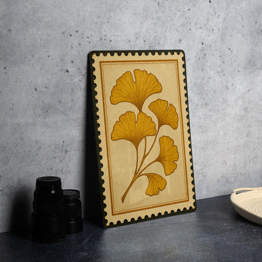 a framed artwork with a yellow ginkgo leaf design, placed on a dark surface next to a stack of black camera lenses.
