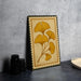 a framed artwork with a yellow ginkgo leaf design, placed on a dark surface next to a stack of black camera lenses.