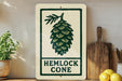 A white sign with a green pine cone graphic and the text "HEMLOCK CONE" is displayed on a wooden shelf, accompanied by a potted plant, a cutting board, and some lemons.