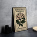 A decorative metal sign with a floral design and text, placed on a table next to some black camera lenses.