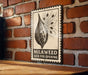 A metal sign with a graphic of a milkweed seed pod and the text "MILKWEED SEED POD OPENING" is displayed on a brick wall.
