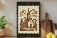 a framed illustration of a church with a steeple, surrounded by trees, on a shelf with various kitchen utensils and a potted plant.