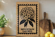 a wooden sign with a floral design, placed on a shelf next to a potted plant and a cutting board.