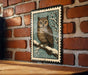 A framed illustration of an owl perched on a branch, set against a snowy background, is displayed on a brick wall.