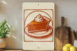 a white sign with an illustration of a slice of pie on a plate, placed on a wooden surface next to a wooden cutting board and a potted plant.