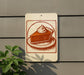 A sign featuring a slice of pie with whipped cream on top, mounted on a wall next to a plant.