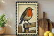A framed illustration of a bird, possibly a robin, perched on a wooden fence. The bird is depicted in a detailed and realistic style, with its vibrant colors and intricate patterns. The frame has a decorative border, adding an artistic touch to the overall presentation. The bird is positioned on a wooden fence, which is a common and recognizable element in outdoor settings.