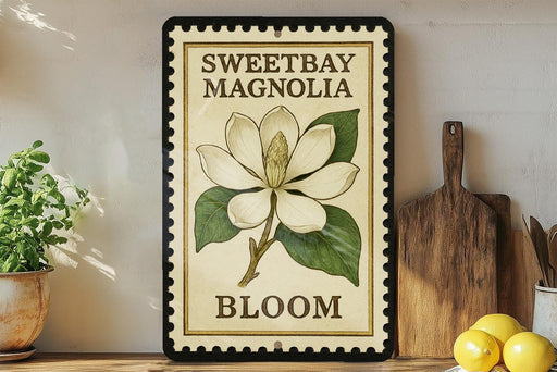 A framed illustration of a white magnolia flower with green leaves, accompanied by the text "SWEETBAY MAGNOLIA BLOOM" on a wooden shelf.