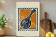 a framed print of a mandolin, a musical instrument, displayed on a shelf next to a wooden cutting board and some lemons.