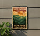 A metal sign with a vintage-style illustration of a mountain landscape, featuring a sun and clouds, mounted on a wall.