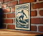 A vintage-style metal sign with a silhouette of a surfer riding a wave, mounted on a brick wall.