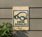 A sign on a wall that says "SWIM GOGGLES" with a graphic of goggles on it.