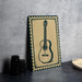 a framed picture of a guitar on a wall, with a stack of black camera lenses and a woven basket placed next to it.