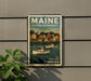 A vintage Maine postage stamp featuring a town scene with boats on the water, mounted on a wall next to a plant.