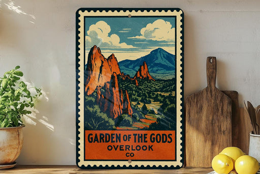 A vintage-style metal sign with a mountain landscape and clouds, featuring the text "GARDEN OF THE GODS OVERLOOK CO" on it. The sign is displayed on a shelf next to a potted plant, a cutting board, and some lemons.
