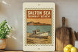 A framed illustration of a boat on a beach, with the text "SALTON SEA BOMBAY BEACH" displayed prominently.
