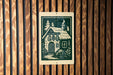A wooden wall with a sign featuring a black and white illustration of a cabin with a chimney, surrounded by trees and bushes.